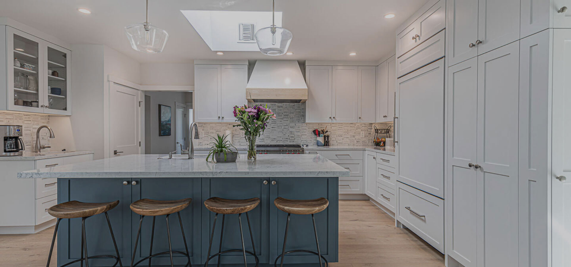 This is an image of a modern kitchen interior with light-colored cabinets, stainless steel appliances, a central island with bar stools, and a large window above the sink area.