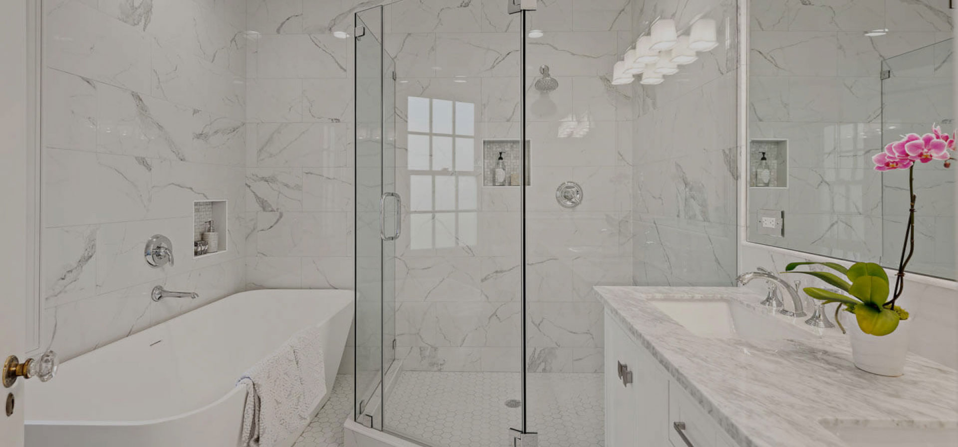 The image shows a modern bathroom interior with a large shower enclosure featuring clear glass doors, a marble-tiled floor, a white toilet, a white sink with a silver faucet, and a vanity mirror above the sink.